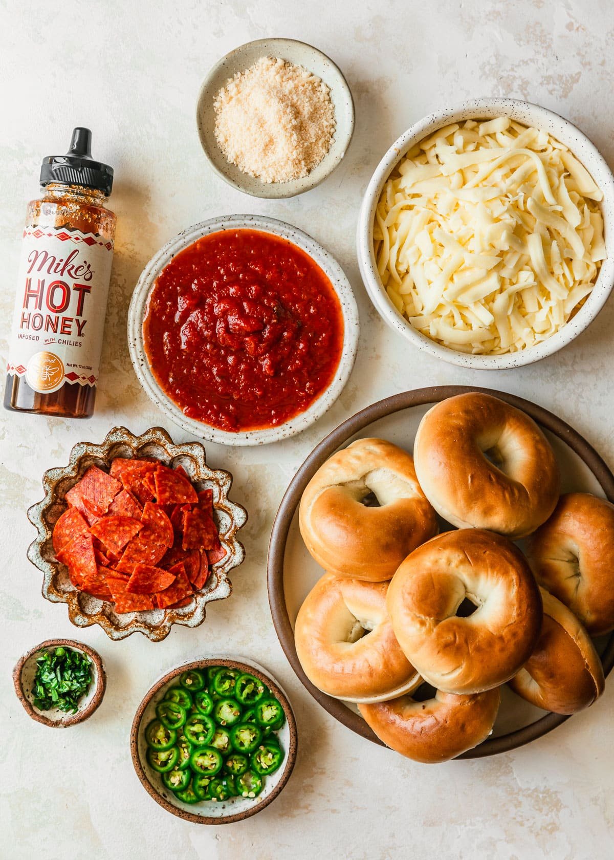 White and brown bowls of bagels, tomato sauce, mozzarella, parmesan, pepperoni, basil, and serrano peppers next to a bottle of hot honey on a tan counter.