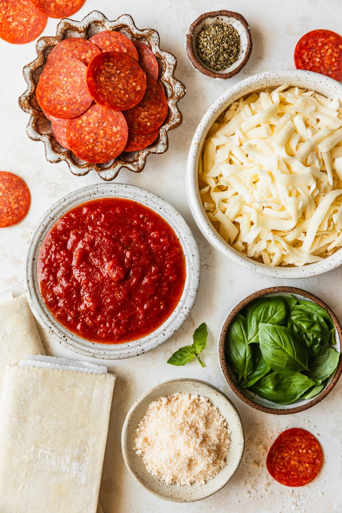 White and brown bowls of red sauce, cheese, pepperoni, basil, and parmesan on a tan counter next to sheets of puff pastry.