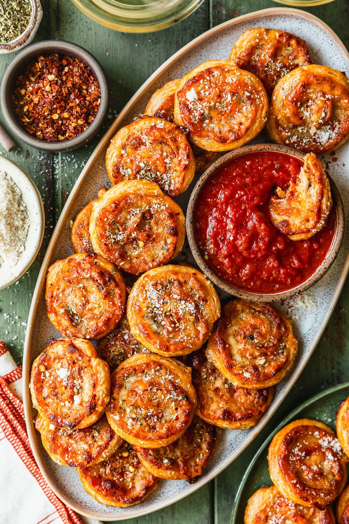 An oval stoneware tray of pizza scrolls on a green wood table next to glasses of white wine and white and brown bowls of oregano, red pepper flakes, and parmesan.