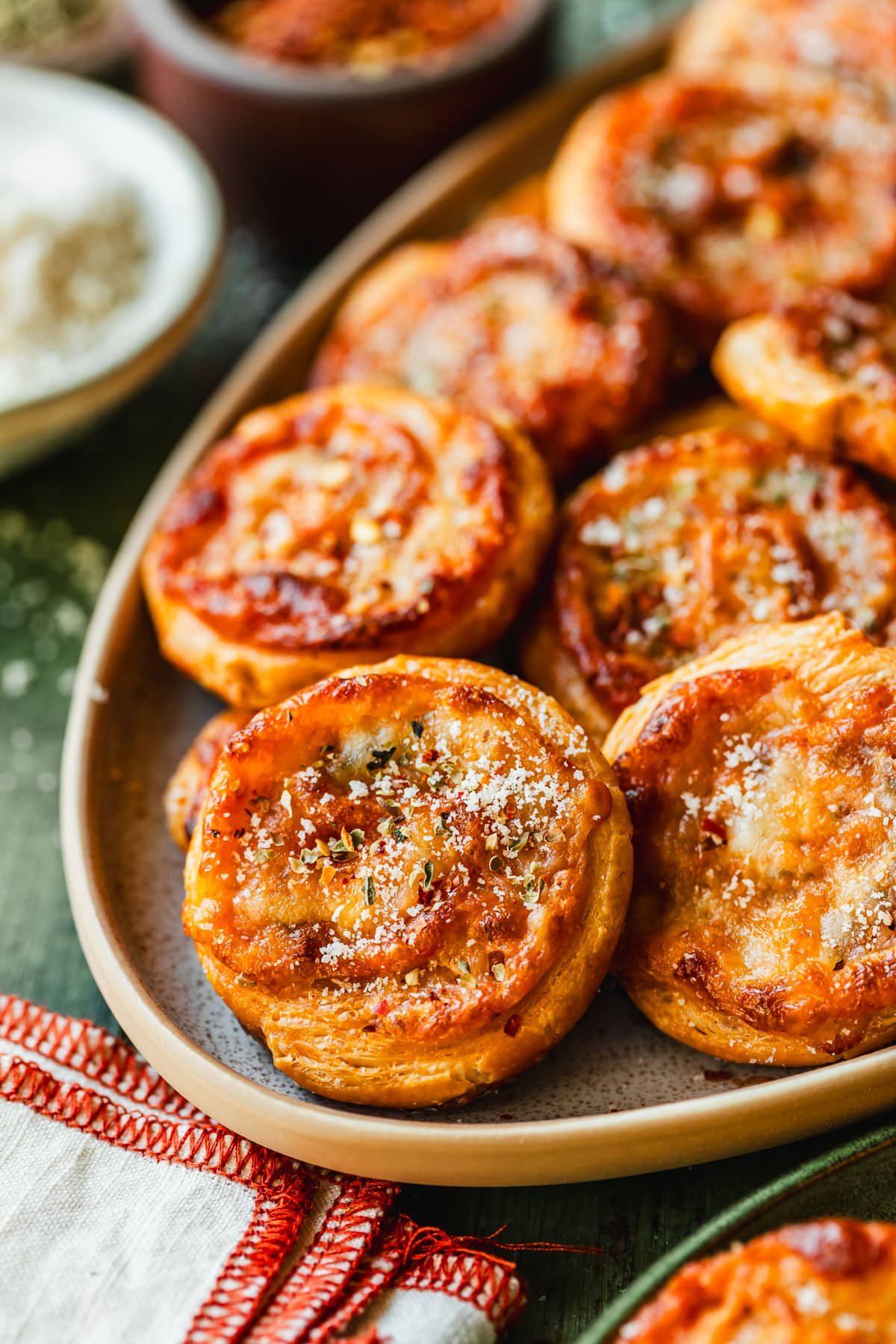 A stoneware platter of pizza scrolls on a green counter next to a green plate, white linen, and white bowl of parmesan.