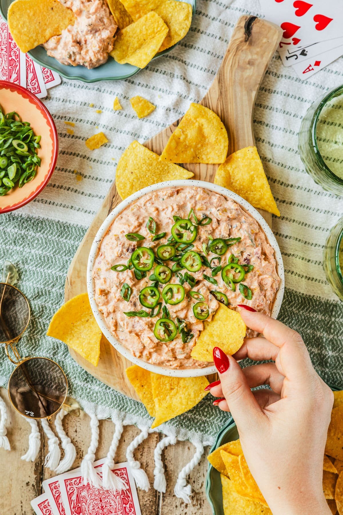 A woman's hand using a corn chip to scoop boat dip out of a white bowl on a striped picnic blanket next to glasses of beer, playing cards, sunglasses, and a blue bowl of corn chips.