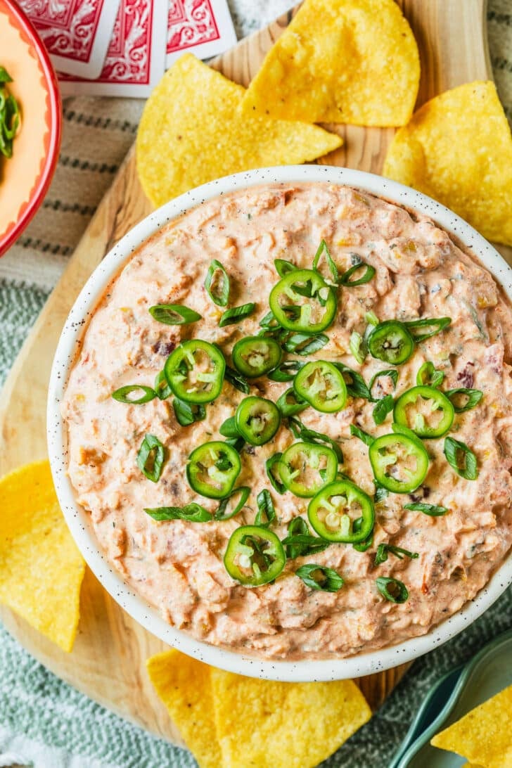 A white bowl of boat dip on a wood board of tortilla chips next to cards and a pink bowl of scallions on a green and white striped blanket.