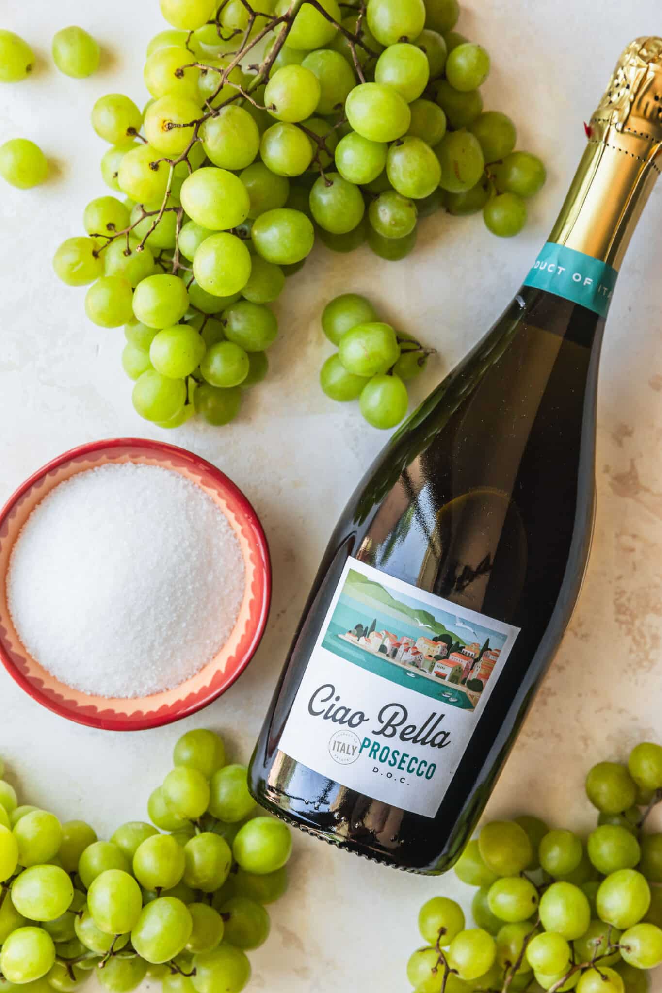 Grapes, a pink bowl of sugar, and a bottle of Prosecco on a tan counter.