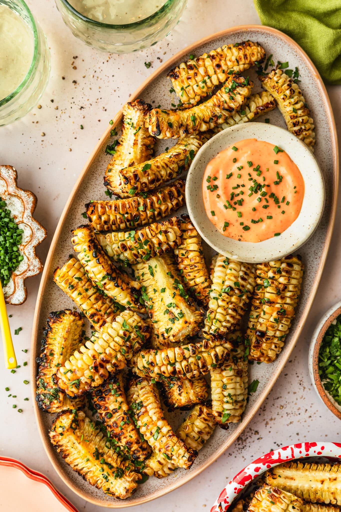 A stoneware platter of grilled corn ribs on a tan counter next to brown bowls of chives and parsley, a red bowl of corn ribs, and a lime green linen.