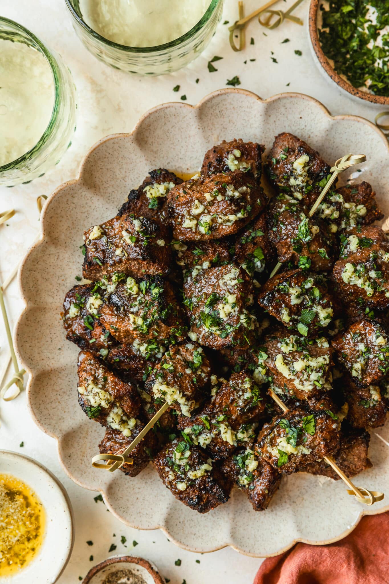 A beige plate of garlic butter steak bites on a tan counter next to an orange linen, glasses of white wine, and a white bowl of garlic butter.