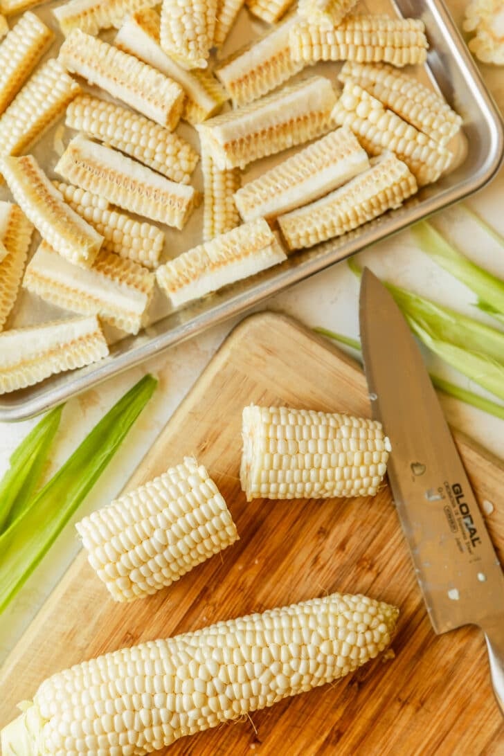 A halved corn cob on a wood board next to a sheet pan of corn.