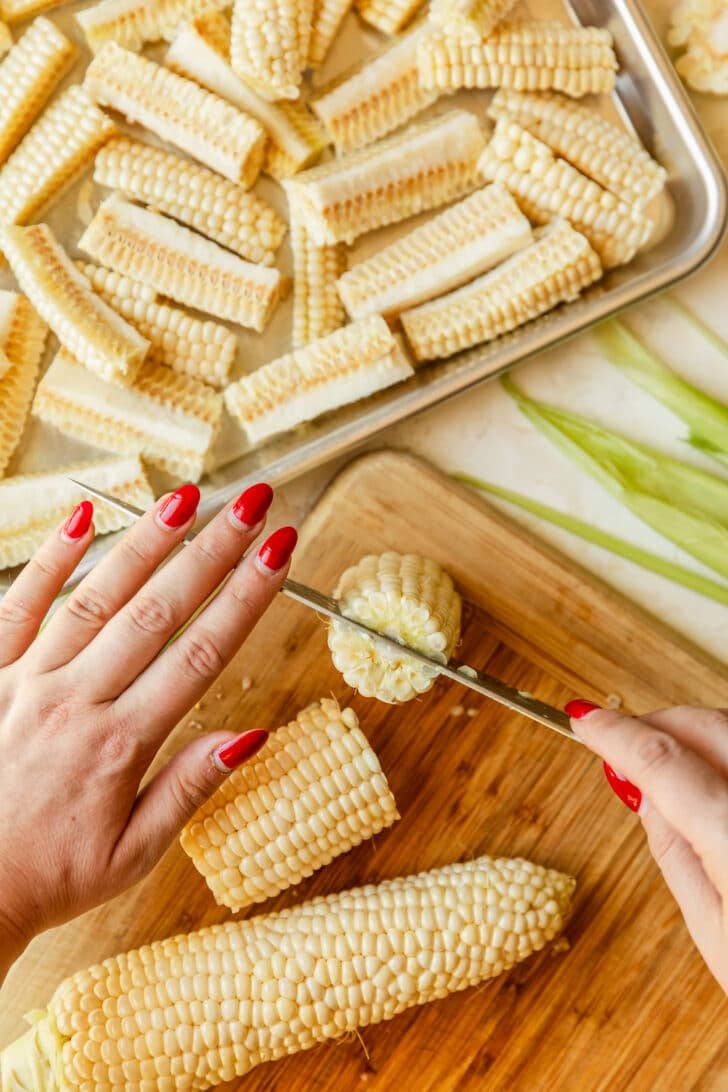 Hands using a knife to cut a corn cob on a wood board.
