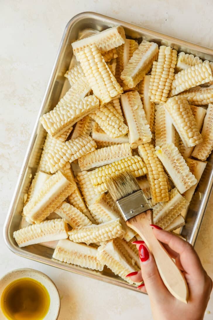 A hand brushing oil on a sheet pan of corn.