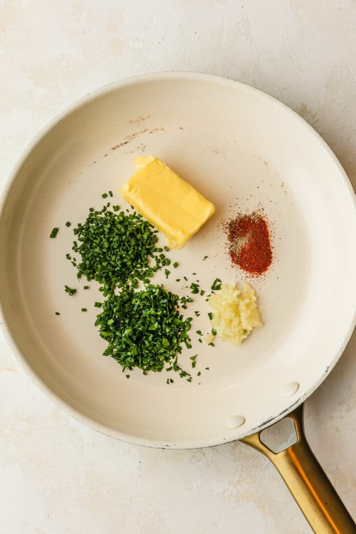 A white pan of garlic, butter, spices, and herbs on a tan counter.