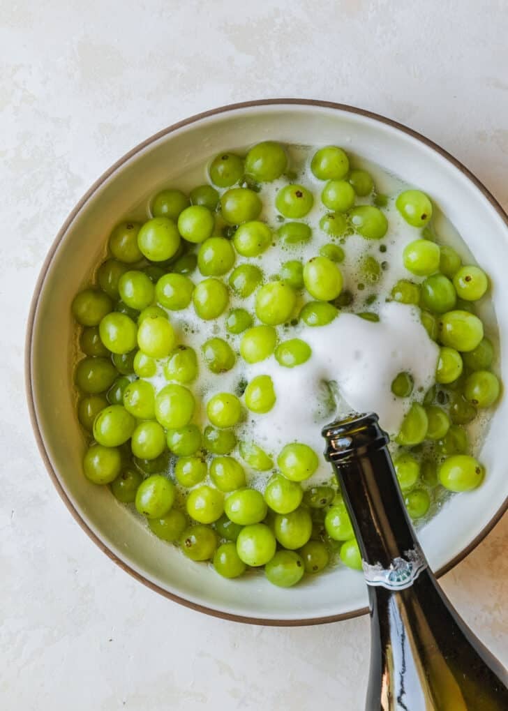 A bottle of bubbly being poured into a white bowl of green fruit.