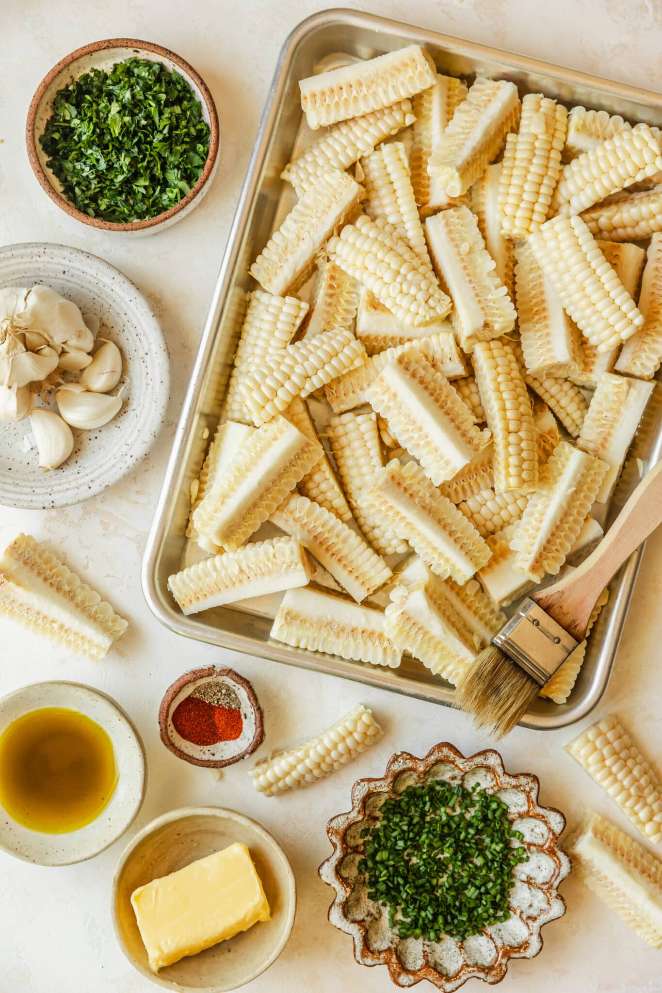 A sheet pan of corn next to brown and white bowls of herbs, garlic, spices, and butter on a tan counter.