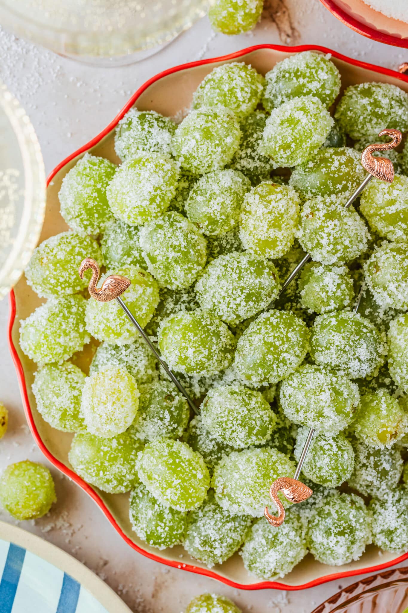 A pink plate of sugared Prosecco grapes on a marble counter next to glasses of Prosecco, a blue striped plate, and a pink bowl of sugar.