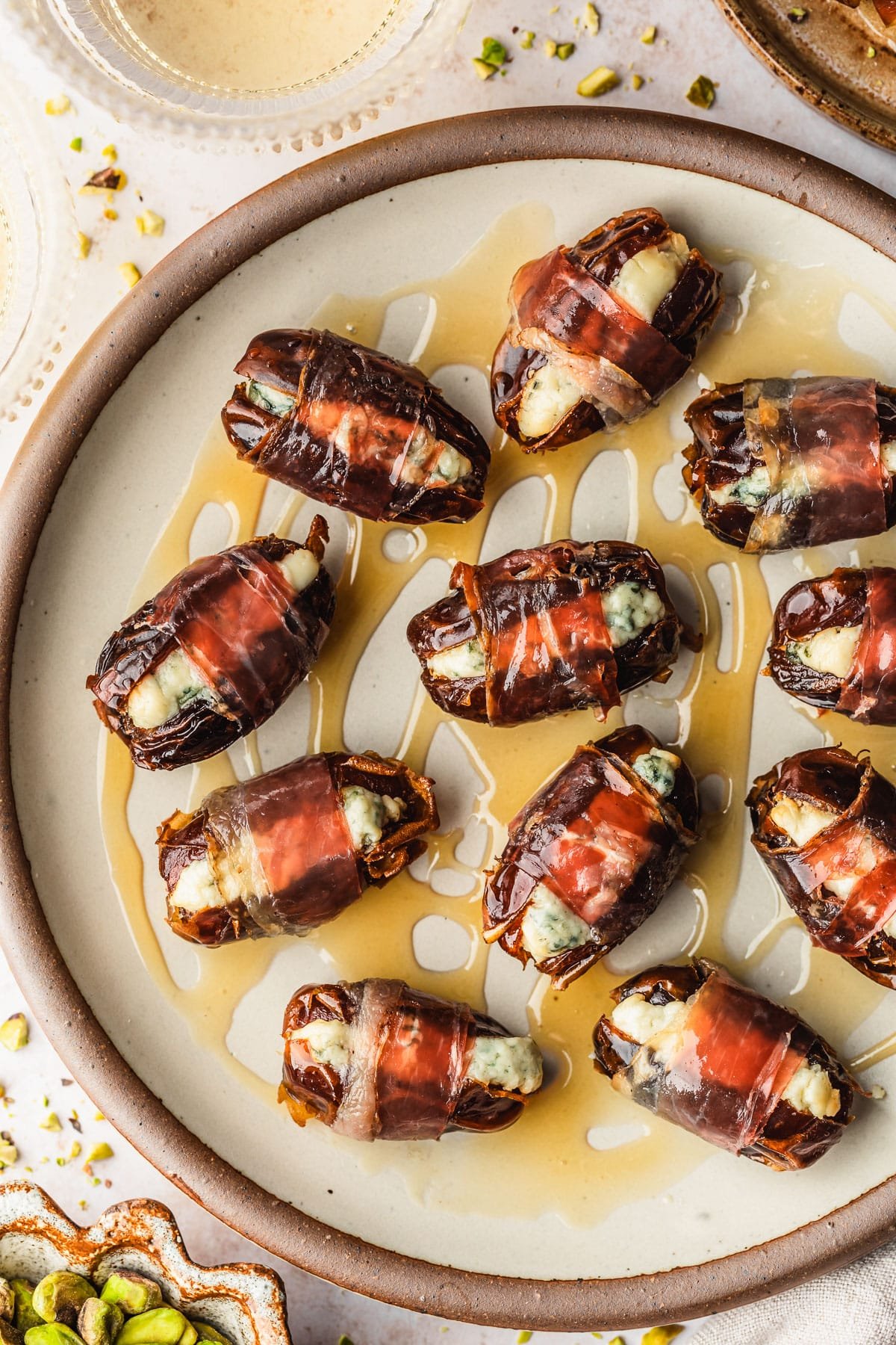 A stoneware plate of blue cheese stuffed prosciutto wrapped dates with honey on a tan counter next to a beige linen, brown bowl of pistachios, and glasses of white wine.