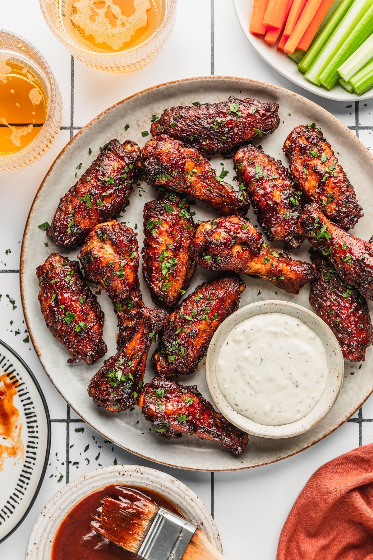 A grey plate of air fryer BBQ chicken wings on a white tiled counter next to an orange linen, glasses of beer, and white bowls of BBQ sauce, carrots, and celery.