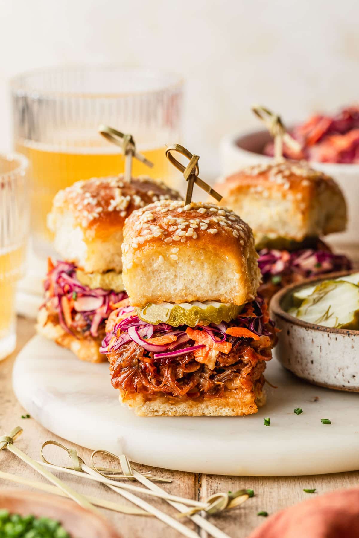 Rows of pulled pork sliders with Hawaiian rolls on a marble platter next to a white bowl of slaw, glasses of beer, and an orange linen with a wood background.
