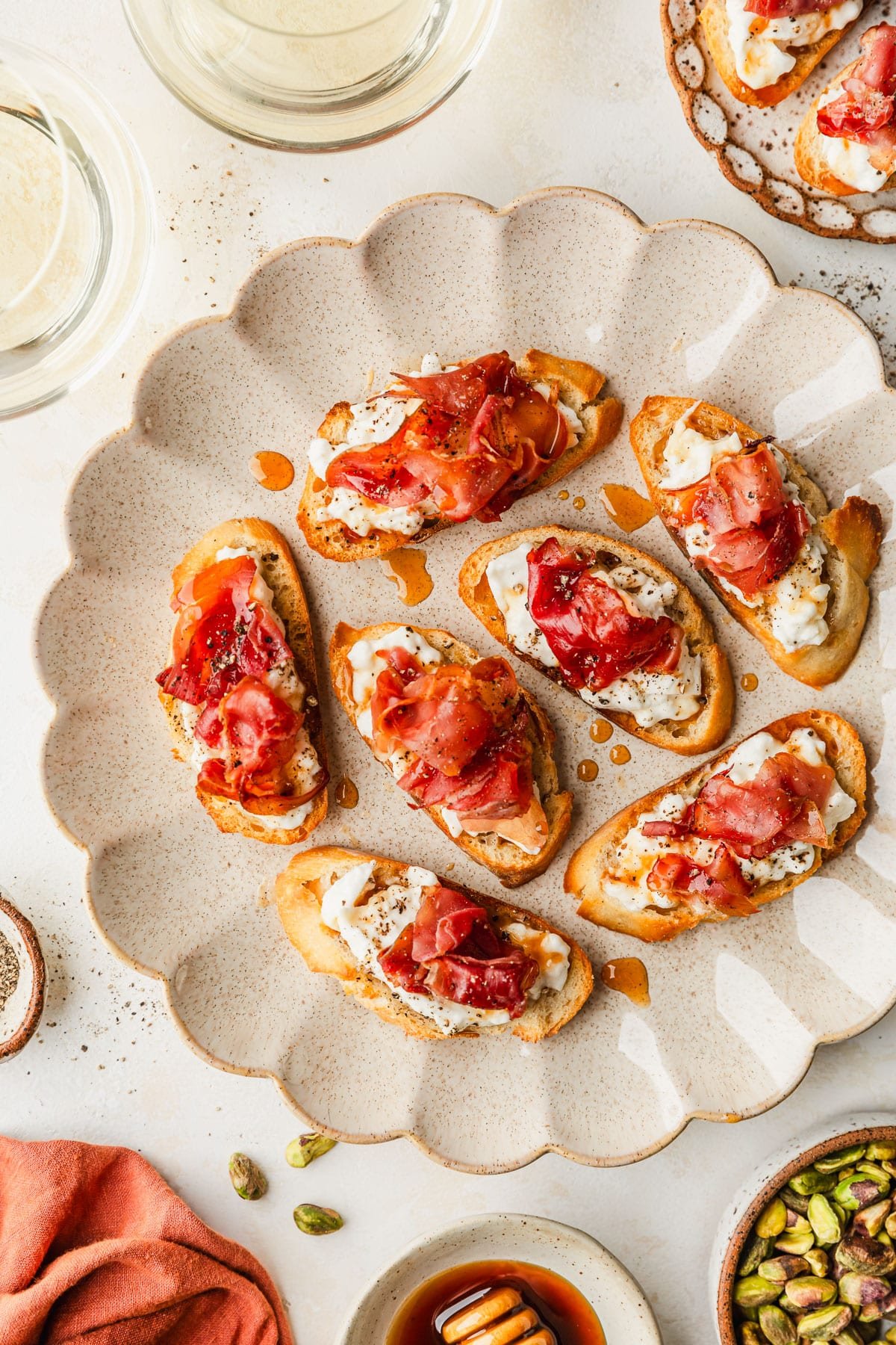 Scalloped plates of prosciutto burrata crostini drizzled with hot honey on a tan counter next to glasses of white wine, an orange linen, a white bowl of hot honey, and a brown bowl of pistachios.