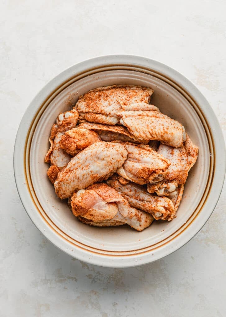 A white bowl of chicken wings tossed in seasoning on a white counter.