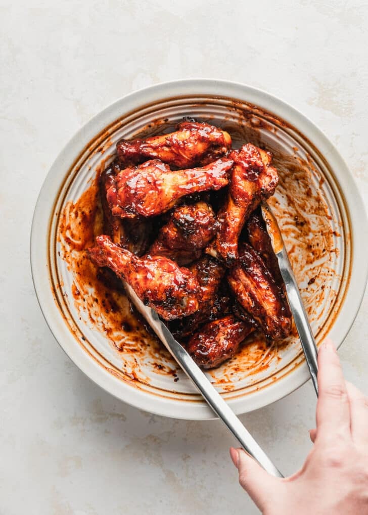 A hand using tongs to toss chicken wings with BBQ sauce in a white bowl.