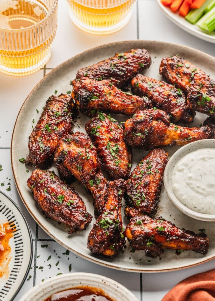 A grey platter of air fryer BBQ chicken wings on a white tiled counter next to glasses of beer, an orange linen, and white bowls of BBQ sauce, celery, and carrots.