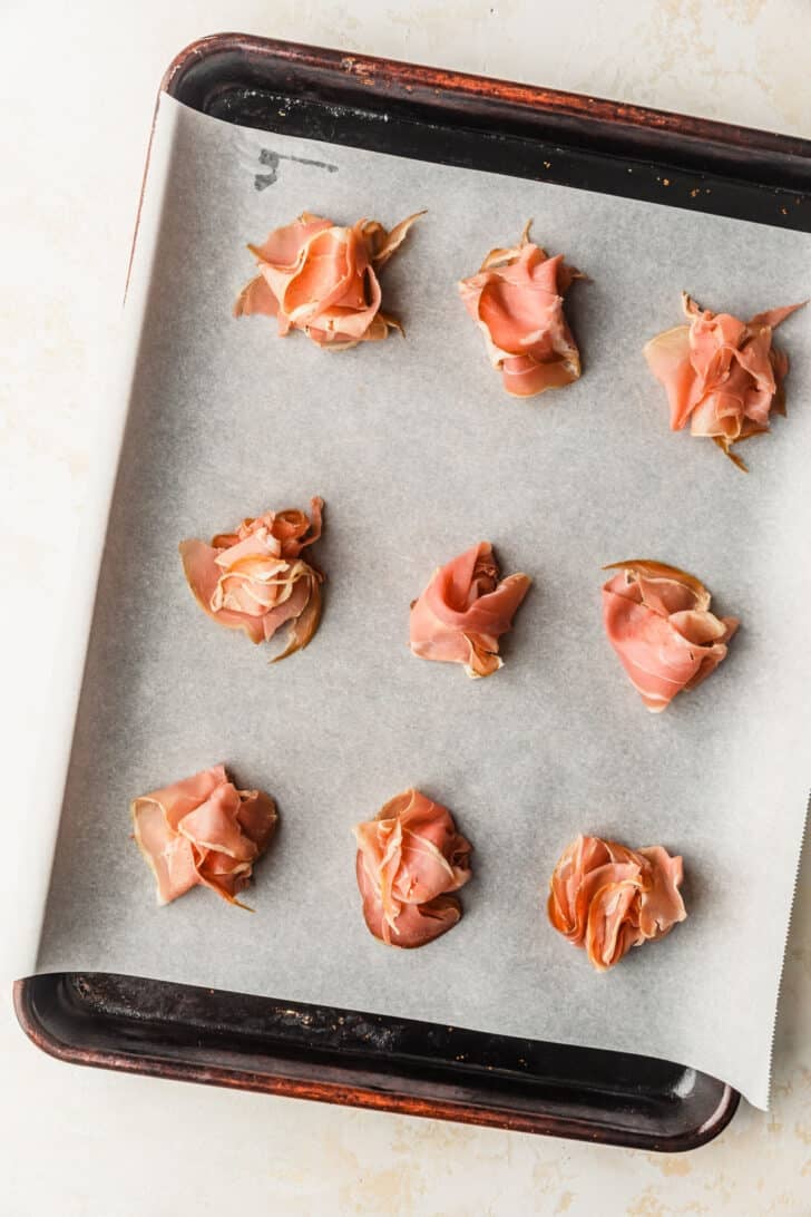 Rows of piled prosciutto on a sheet pan with a tan background.