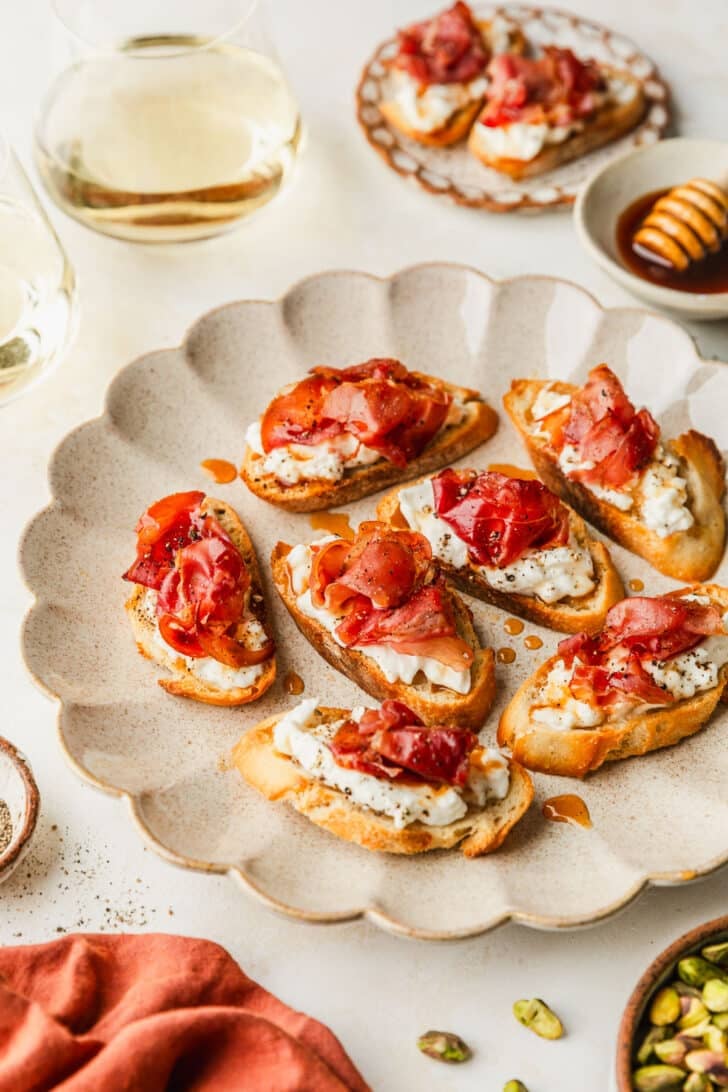 A tan scalloped plate of burrata and prosciutto crostini on a beige table next to an orange linen, white bowl of honey, glasses of wine, and brown bowl of pistachios.