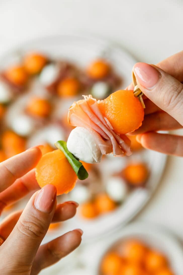 Hands skewering melon, prosciutto, mozzarella, and basil on skewers.