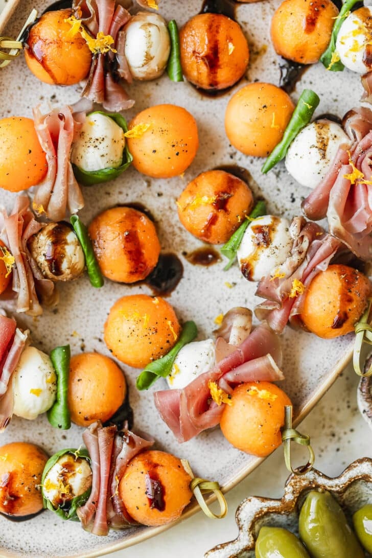 A stoneware plate of prosciutto melon skewers with balsamic glaze on a white counter next to a brown and white bowl of olives.