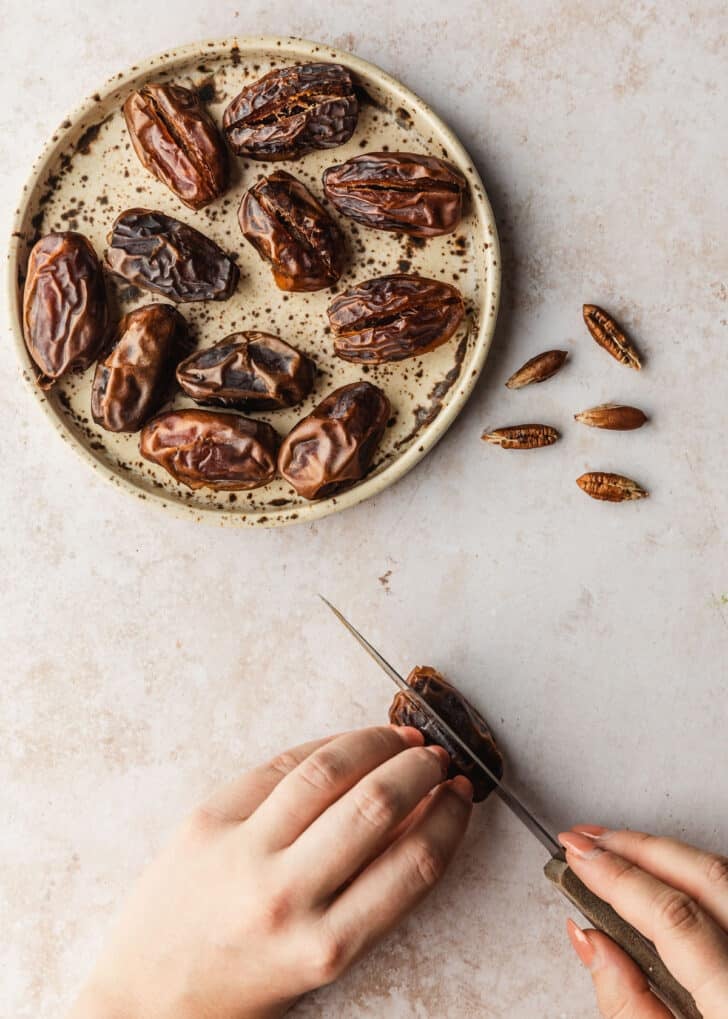 A hand using a knife to slice open a date on a tan counter next to a brown plate of dates.