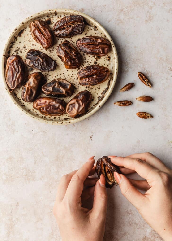Hands opening up a date on a tan counter next to a brown plate of dates.