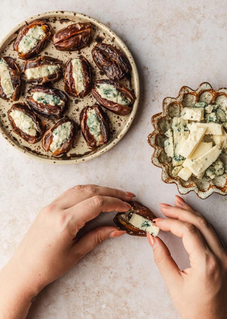Hands placing a wedge of blue cheese into a date next to brown bowls of dates and blue cheese on a tan counter.
