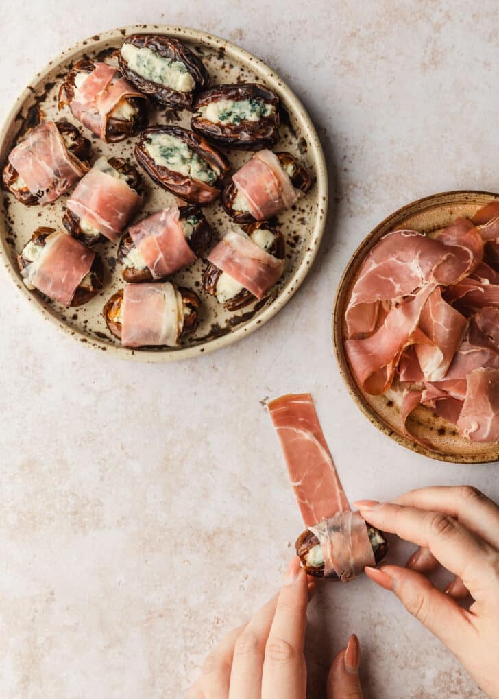 Hands rolling dates in prosciutto on a tan counter.