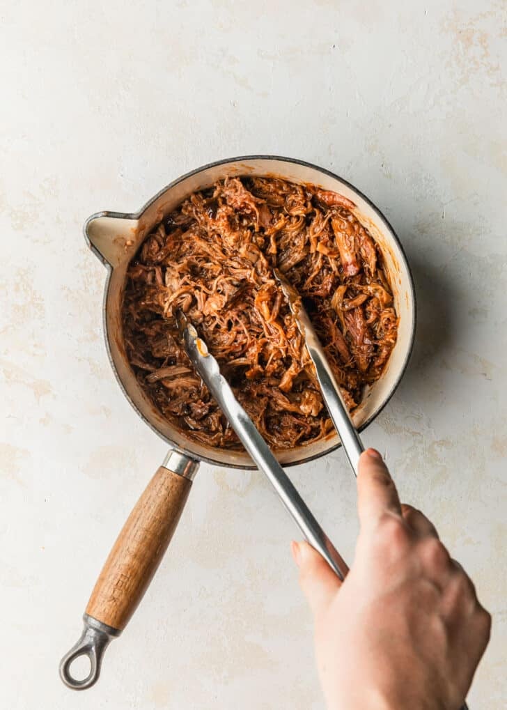Hands using tongs to toss pulled pork and BBQ sauce in a small pot.