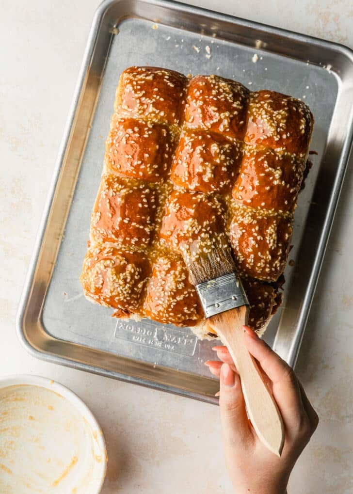 A hand using a pastry brush to brush butter mixture on the top of rolls.