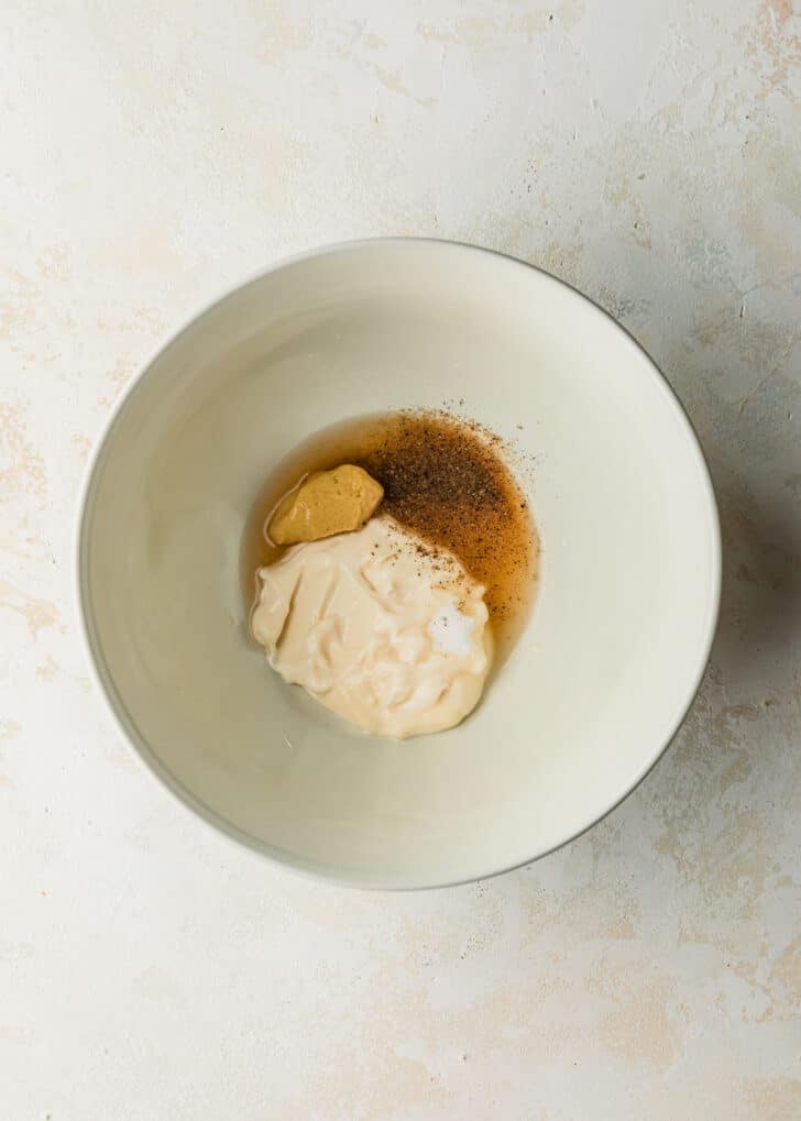 A white bowl of mayo, vinegar, mustard, & spices on a beige counter.