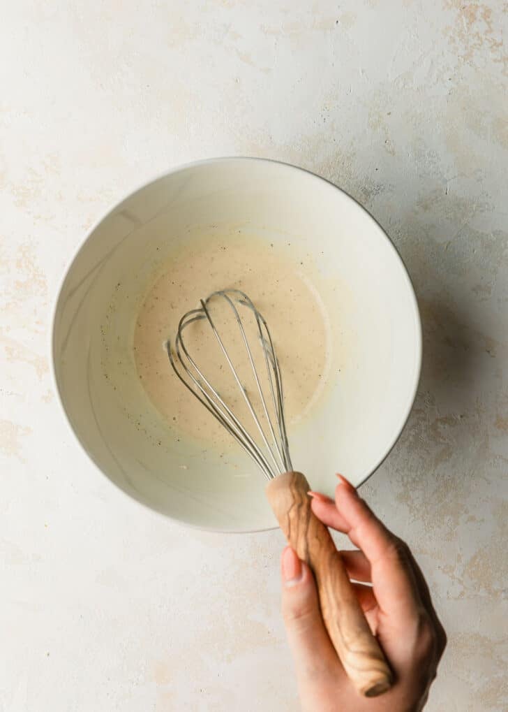 A hand using a whisk to mix dressing in a white bowl.