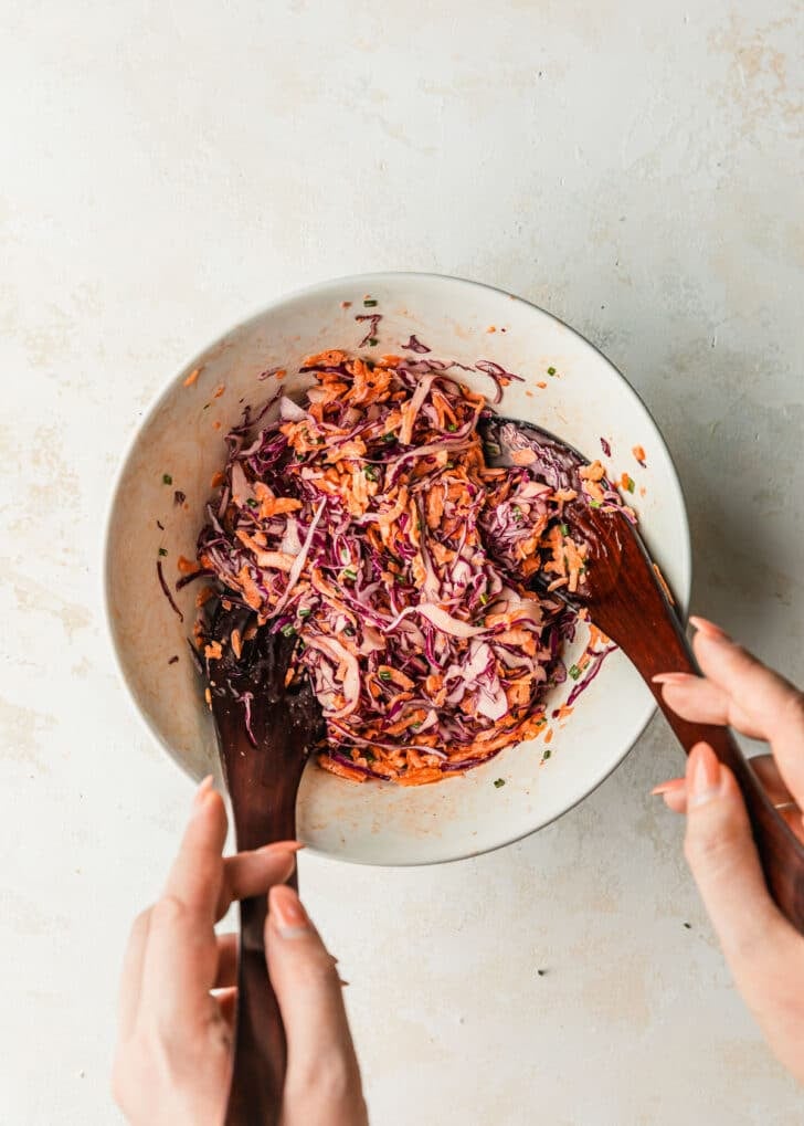 Hands using salad tongs to toss coleslaw for pulled pork in a white bowl.