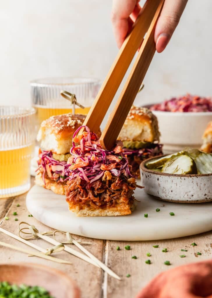 A hand using tongs to place coleslaw on a white tray pulled pork sliders. The tray is on a wood counter surrounded by a white bowl of slaw, glasses of beer, and an orange towel.