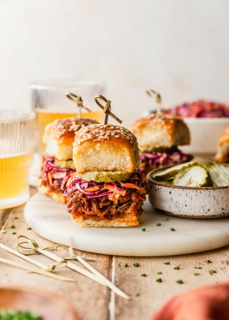 A white tray of pulled pork sliders with Hawaiian rolls and coleslaw on a wood table next to glasses of beer, a white bowl of slaw, and an orange napkin.