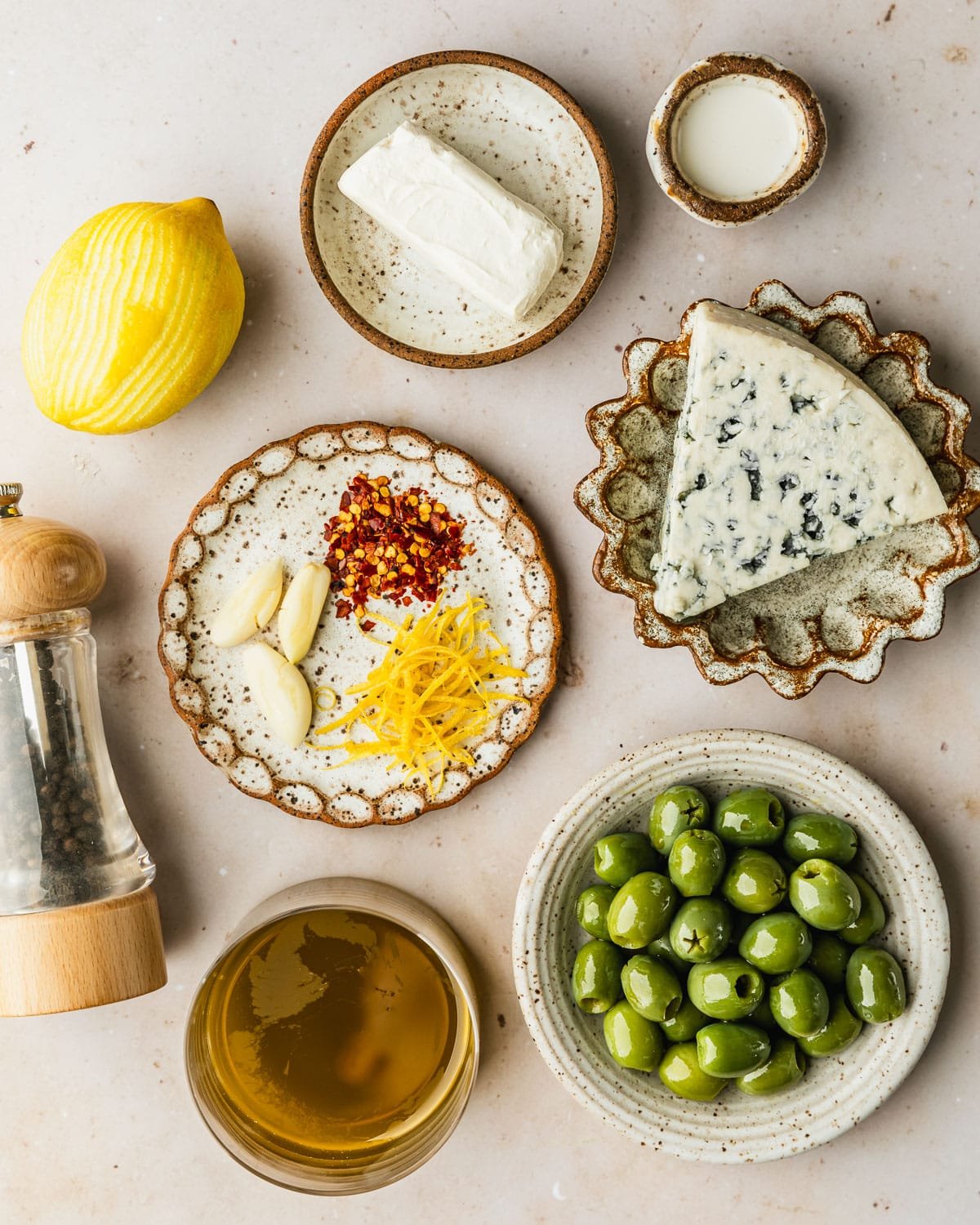 Brown and white bowls of olives, blue cheese, cream cheese, cream, lemon zest, pepper flakes, garlic, and olive oil next to a pepper grinder and lemon on a tan counter.