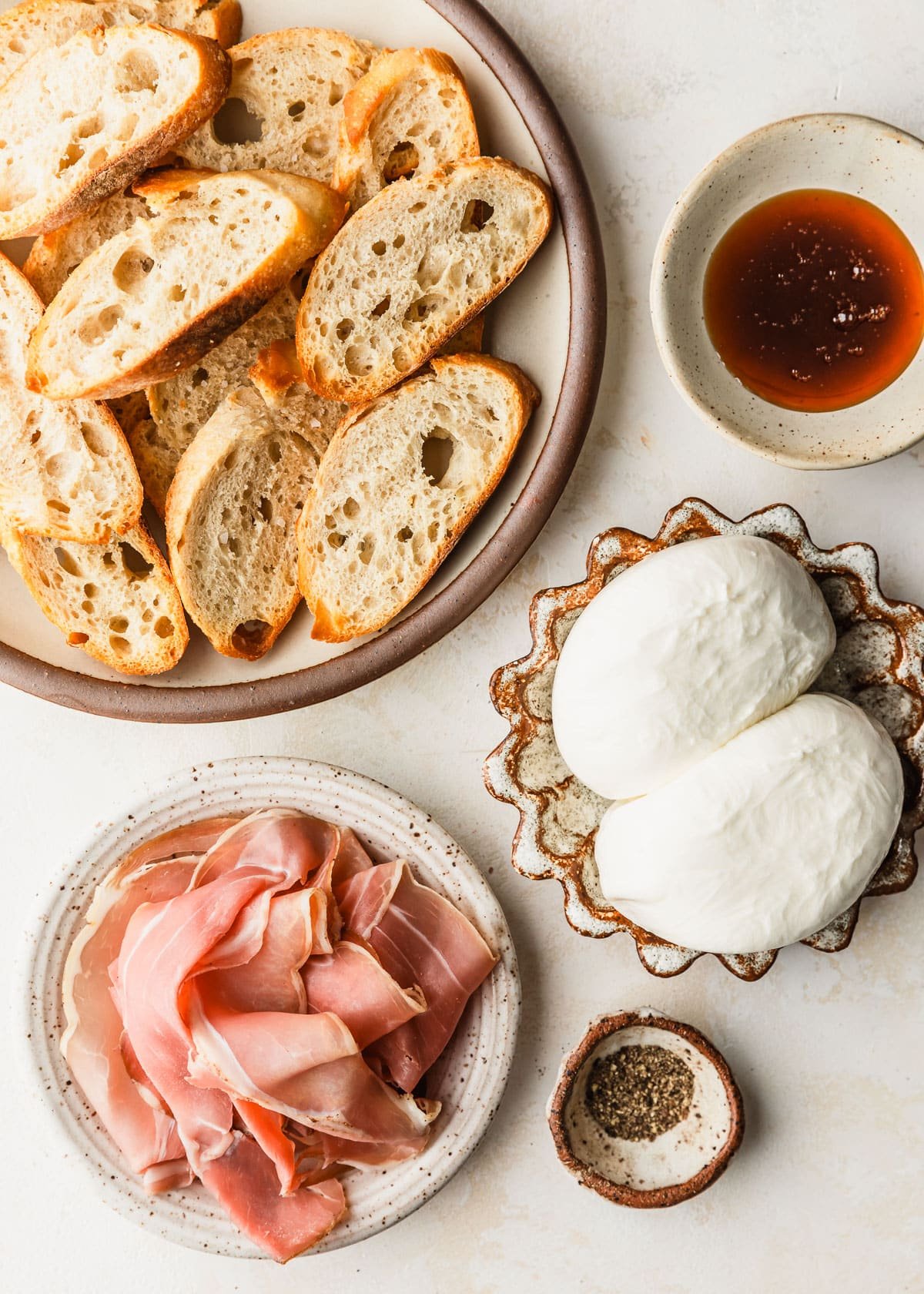 White and brown bowls of bread, mozzarella, deli meat, black pepper, and hot honey on a beige counter.