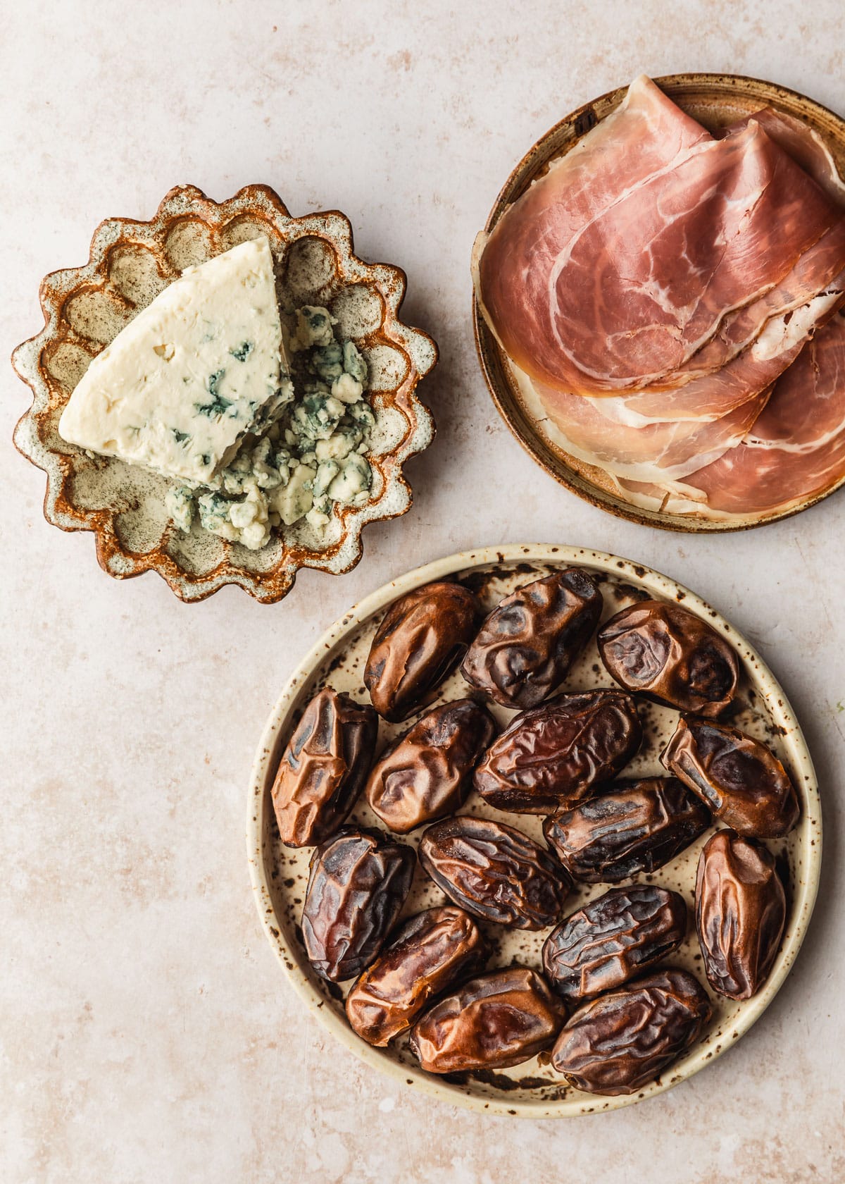Brown plates of blue cheese, dates, and prosciutto on a tan counter.
