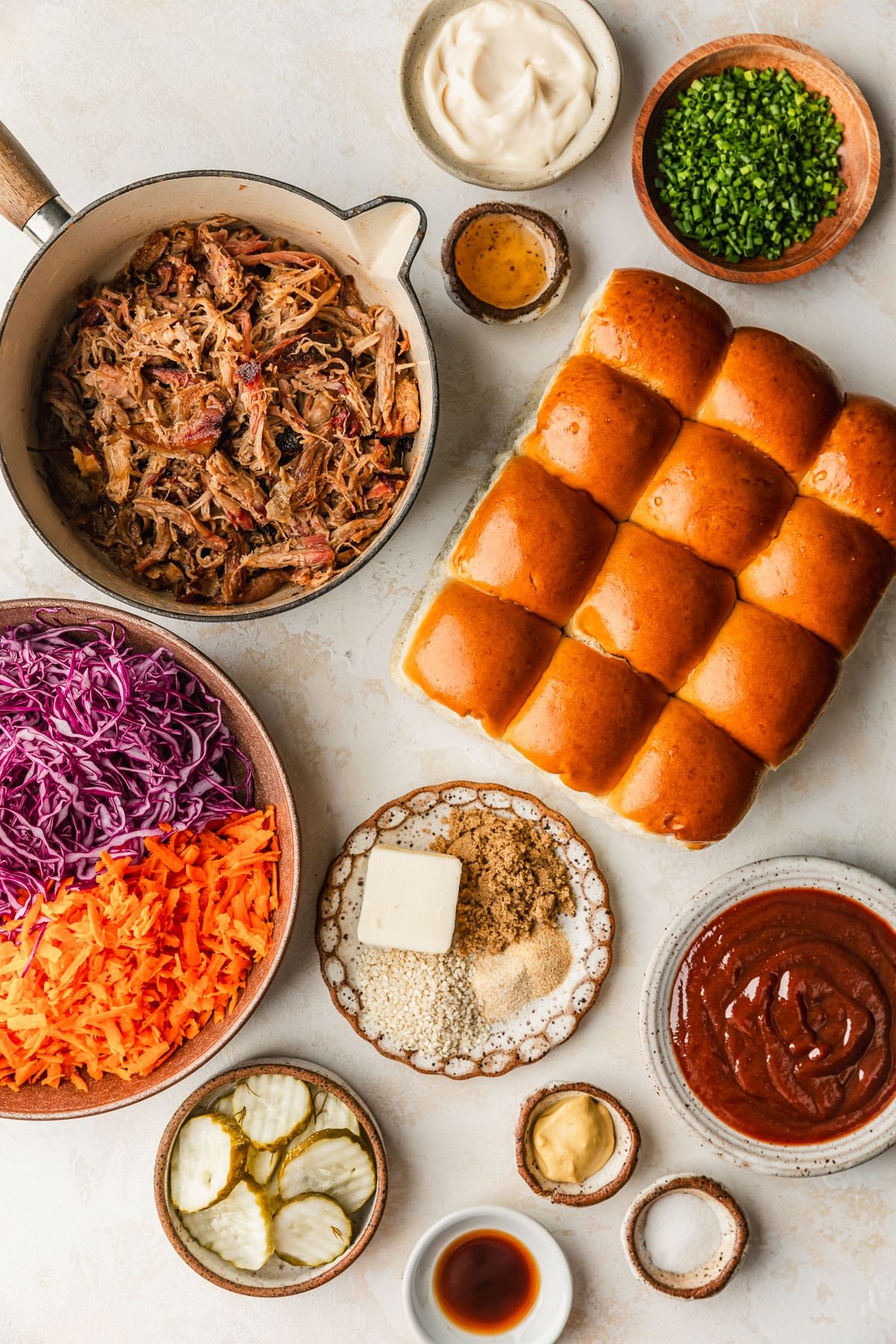White and brown bowls of shredded meat, rolls, mayo, chives, vinegar, cabbage, carrots, spices, BBQ sauce, pickles, mustard, sugar, and Worcestershire sauce on a tan counter.