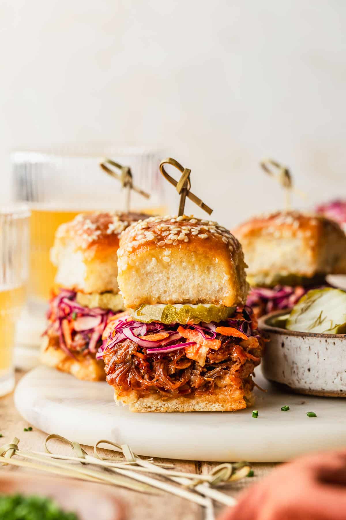 A white platter of BBQ pulled pork sliders with Hawaiian rolls on a wood table next to glasses of beer, an orange linen, and a white bowl of pickles.