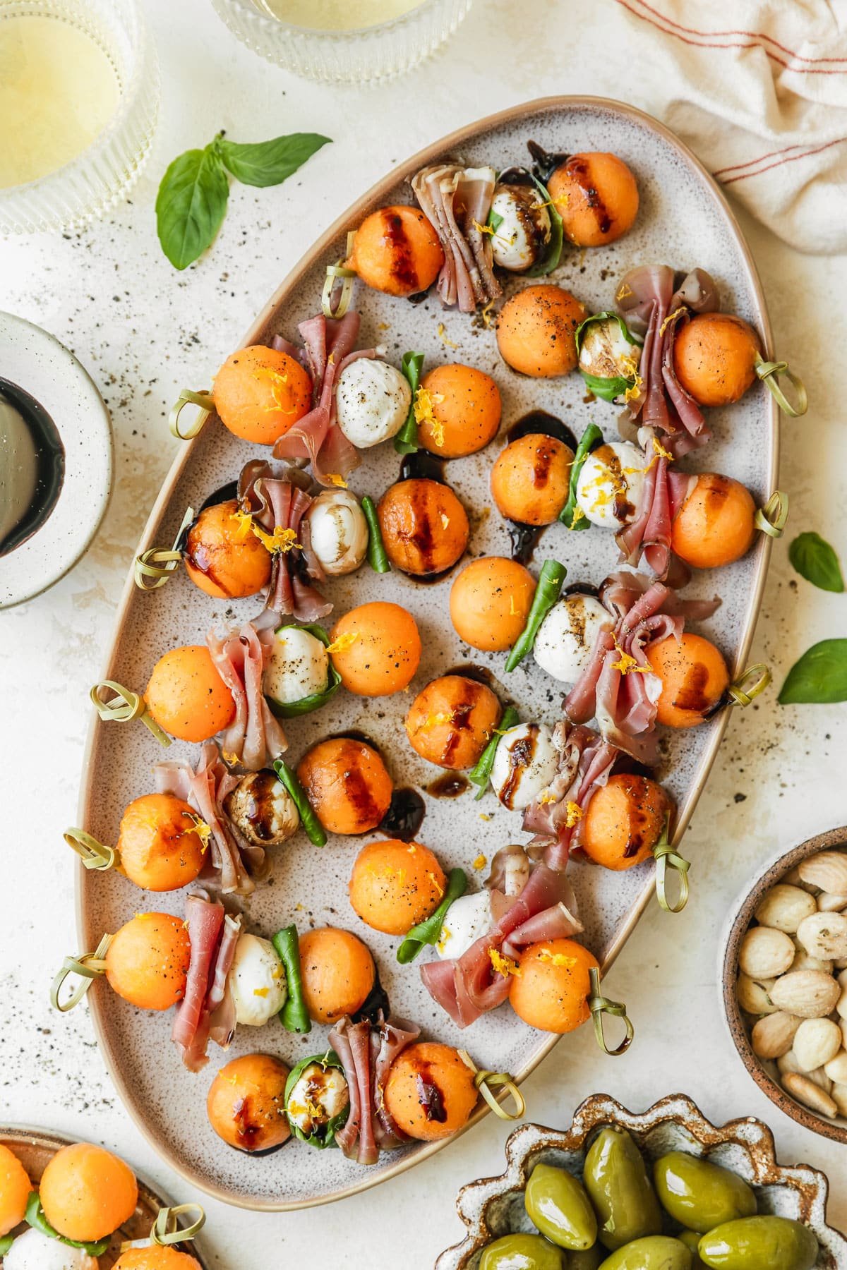 A stoneware platter of melon prosciutto skewers on a white counter next to glasses of white wine, a cream-colored linen, a white bowl of balsamic glaze, and brown bowls of olives and almonds.