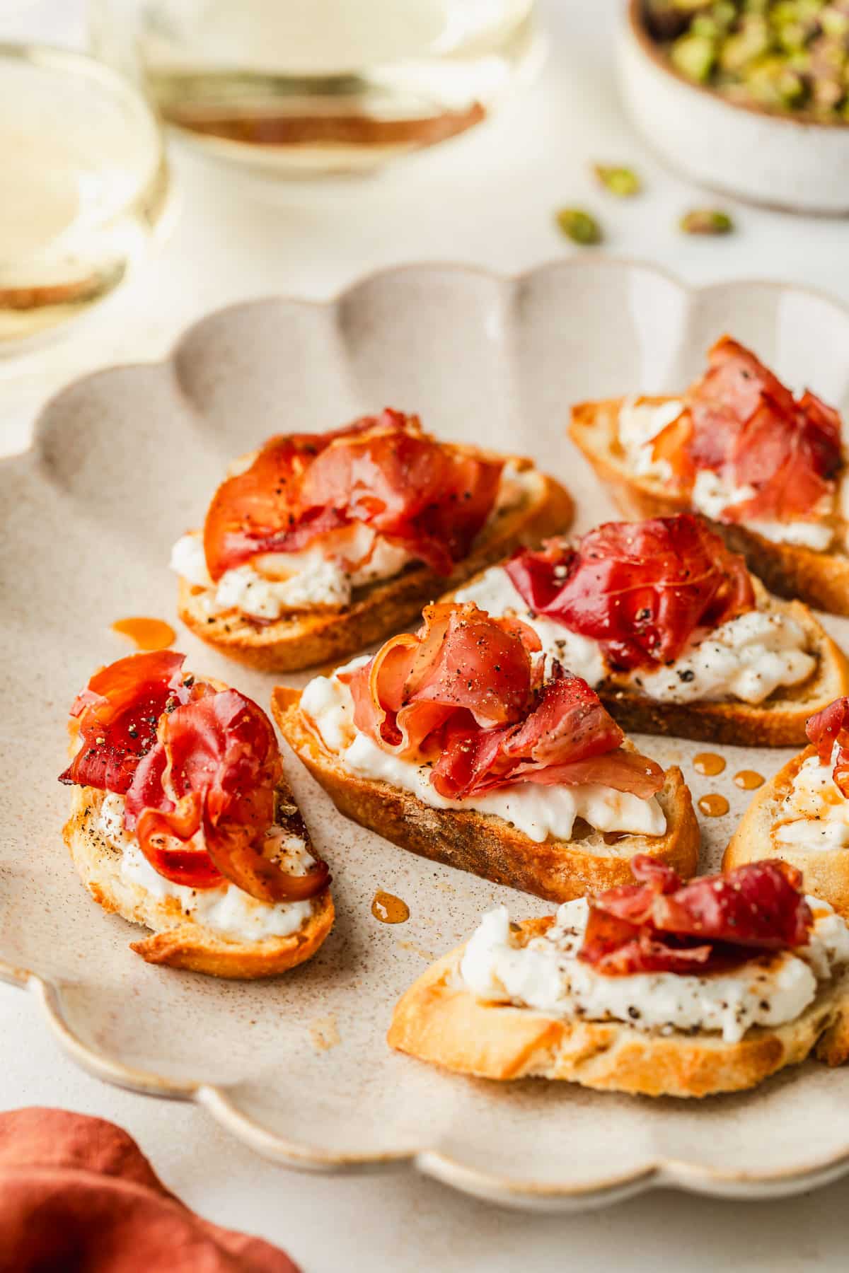 A beige plate of burrata prosciutto crostini on a tan counter next to an orange linen, glasses of white wine, and a white bowl of pistachios.