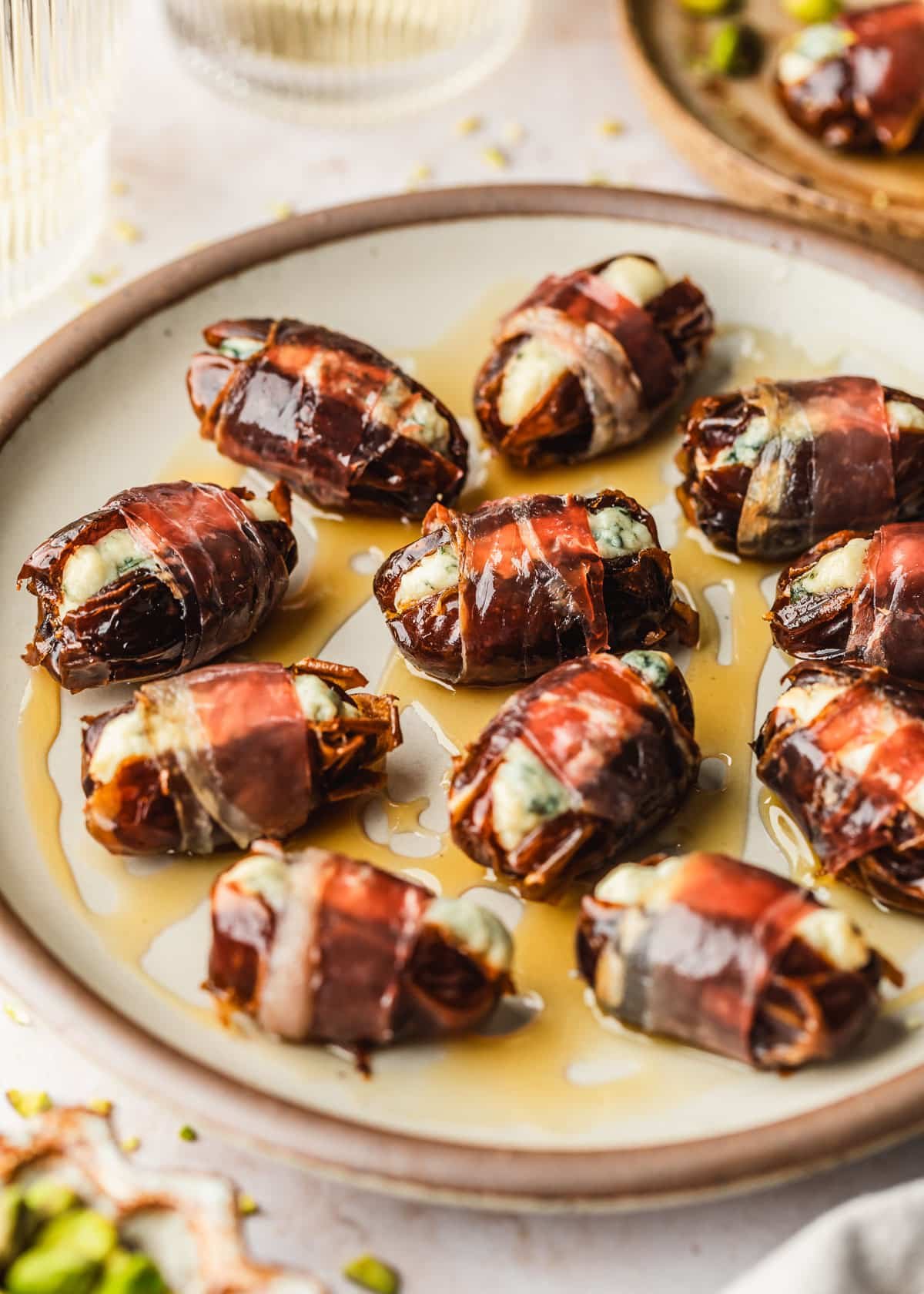 A stoneware plate with honey and blue cheese stuffed prosciutto wrapped dates on a tan counter next to glasses of white wine, a brown bowl of pistachios, and a brown plate.