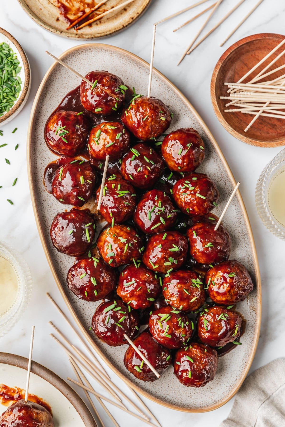 A stoneware platter of BBQ grape jelly meatballs on a white marble counter next to glasses of white wine, a brown linen, and white and brown bowls of chives and toothpicks.