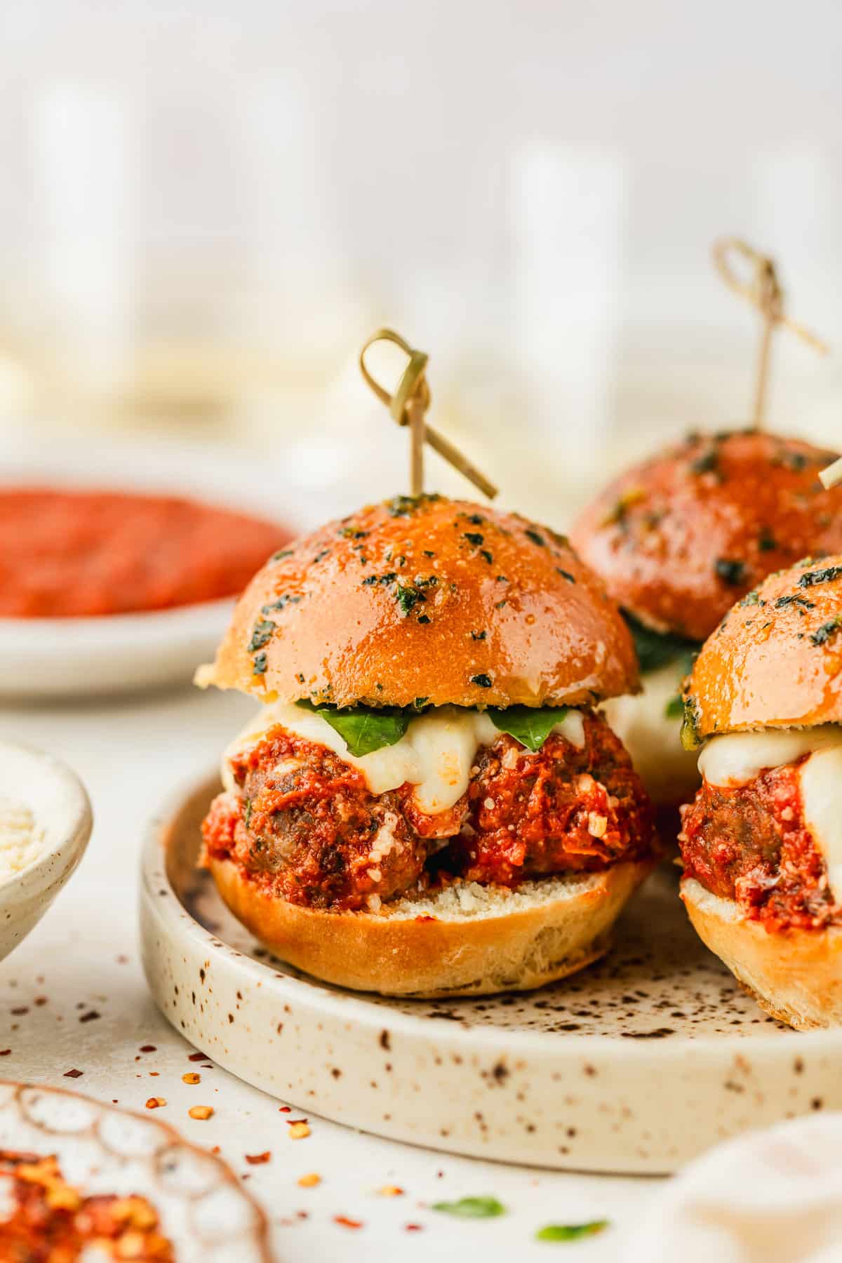 A speckled plate with Italian meatball sliders on a tan counter next to white bowls of parmesan, red sauce, and pepper flakes.
