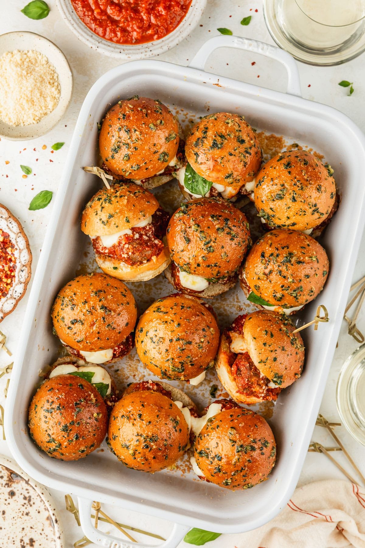 A white pan of Italian meatball sliders next to glasses of white wine, a cream-colored linen, and white bowls of tomato sauce, parmesan, and red pepper flakes.