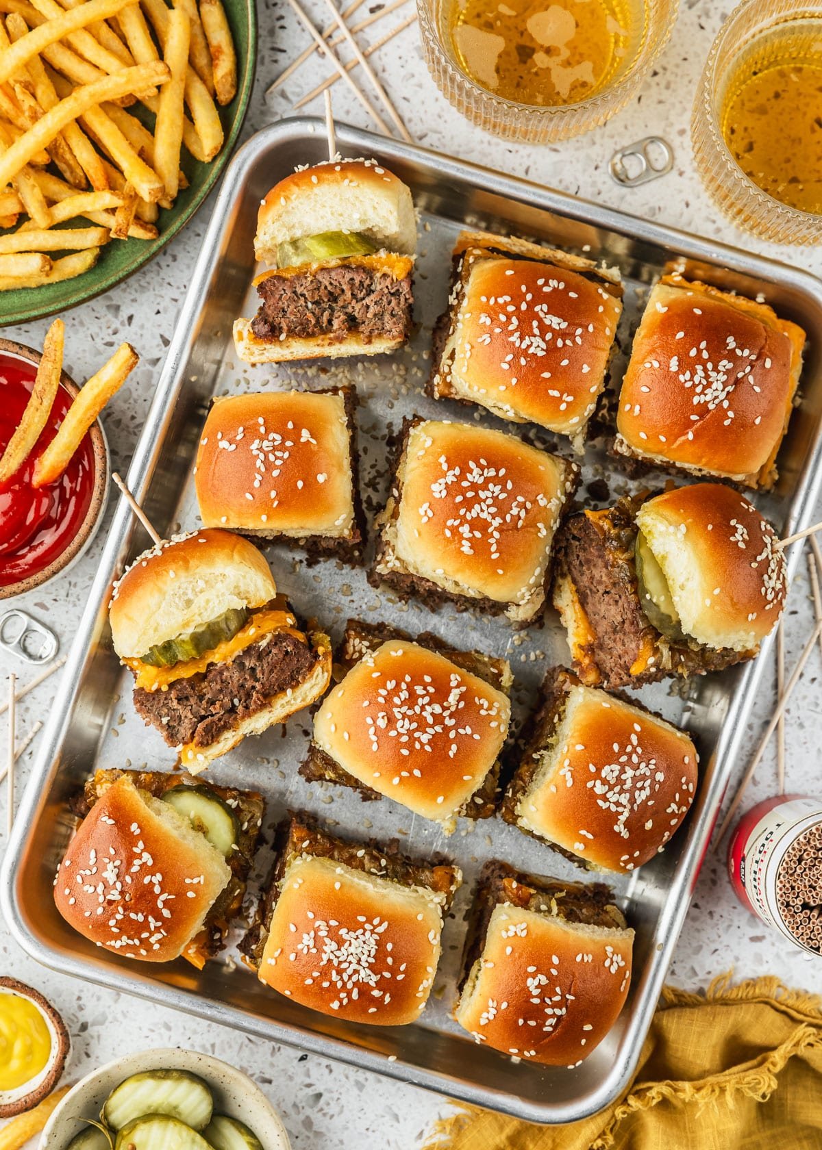 A sheet pan of cheeseburger sliders with Hawaiian rolls on a white terrazzo counter next to a green plate of fries, glasses of beer, and white bowls of ketchup, mustard, and pickles.